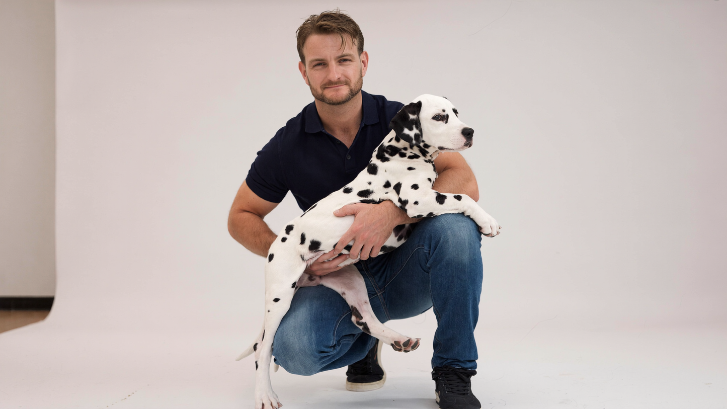 A man crouching in a studio while holding a Dalmatian puppy. The man is wearing a dark navy polo shirt and blue jeans, and the puppy is white with black spots.