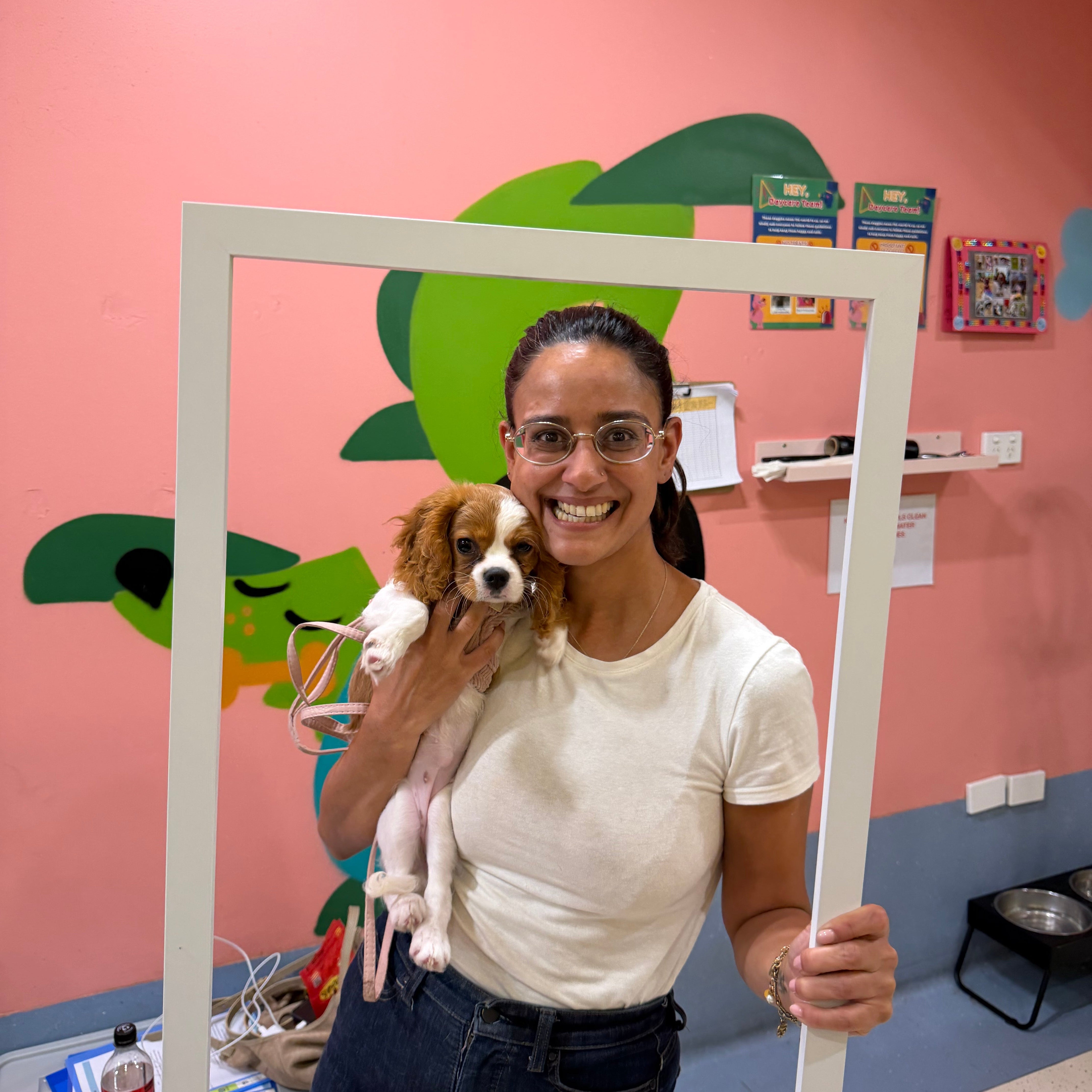 A smiling woman wearing glasses and a white t-shirt holds a small brown and white dog inside a large white square frame, set against a pink wall with green cartoon graphics.