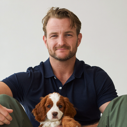 A man sitting on the floor wearing a navy polo shirt and olive green pants, holding a brown and white spaniel puppy against a plain light gray background.