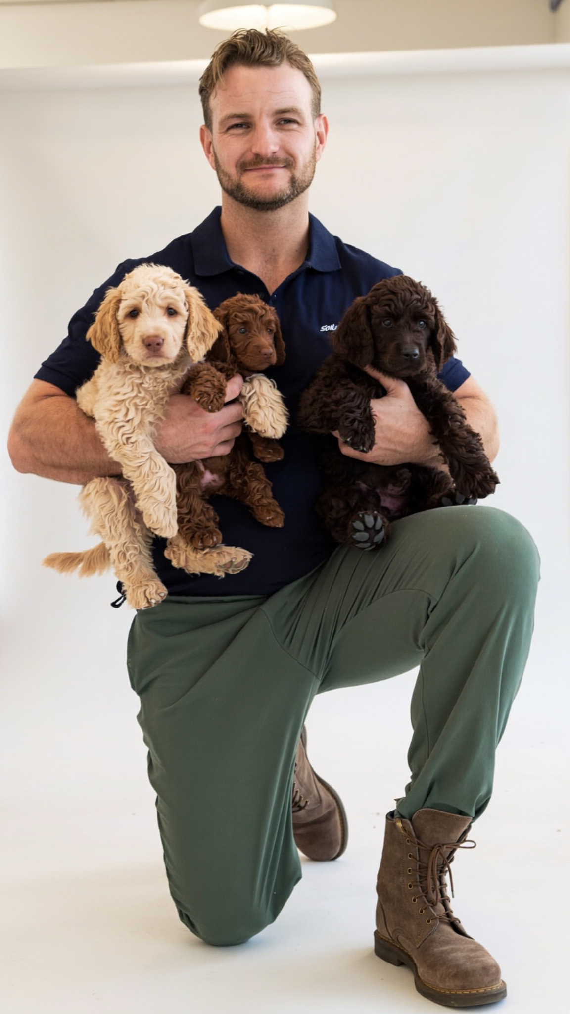 A man kneeling in a white studio setting, holding three puppies. He is wearing a navy blue polo shirt, olive green pants, and brown lace-up boots.