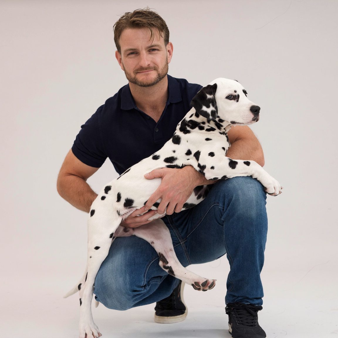 A man crouching in a studio while holding a Dalmatian puppy. The man is wearing a dark navy polo shirt and blue jeans, and the puppy is white with black spots.