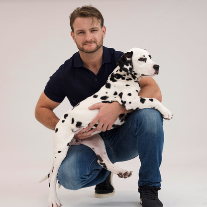 A man crouching in a studio while holding a Dalmatian puppy. The man is wearing a dark navy polo shirt and blue jeans, and the puppy is white with black spots.