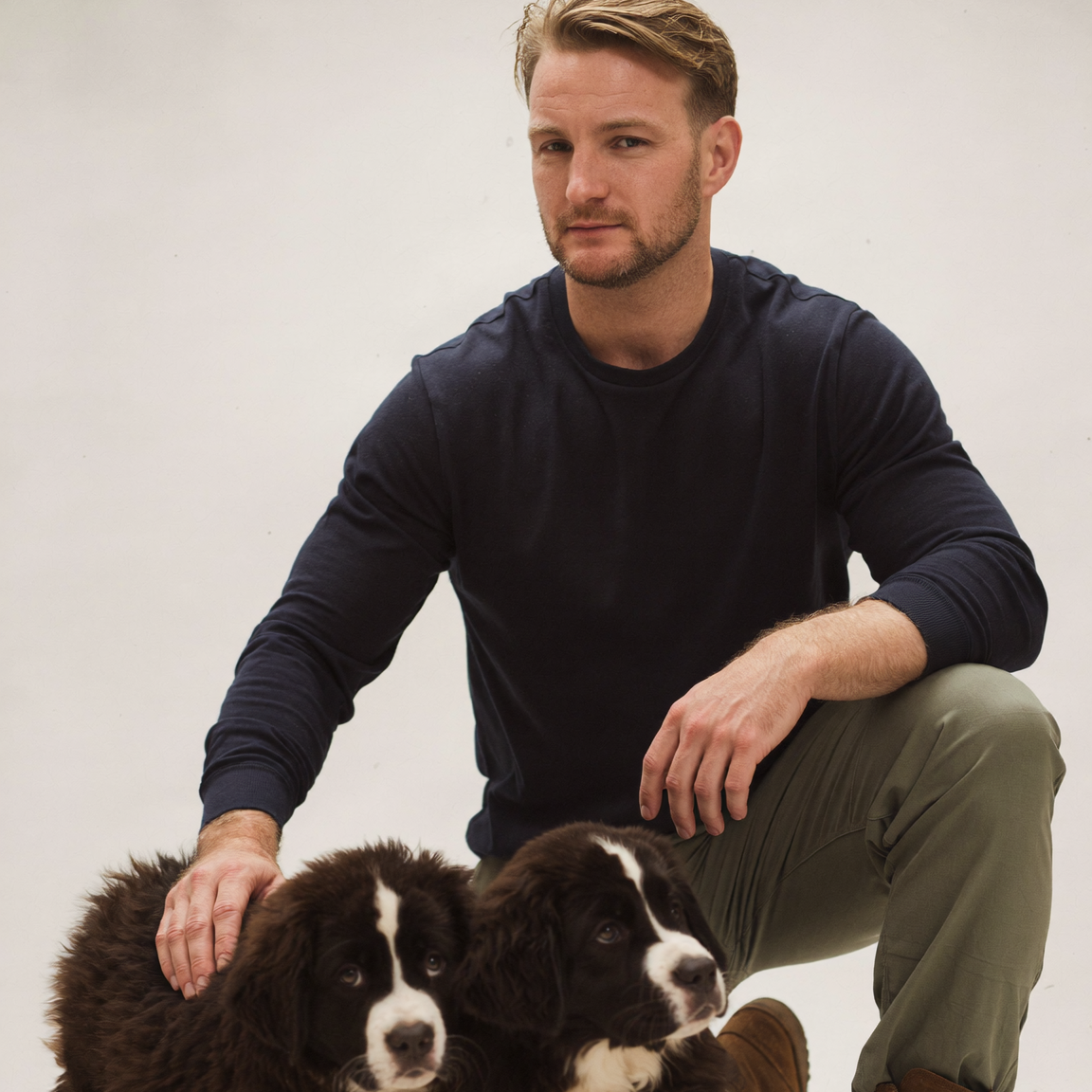 A man crouching on a light studio floor with two black-and-white puppies. He is wearing a dark navy long-sleeve shirt, olive green pants, and brown boots.